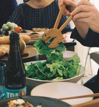 Load image into Gallery viewer, A hand carved pair of salad servers picking up a green salad at a dining table full of people and food.