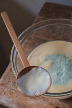 Load image into Gallery viewer, A hand carved spoon resting on the side of a glass bowl with a wet mixture in it.