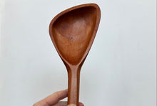 Load image into Gallery viewer, A hand holding a large wooden serving spoon with a smooth, rounded bowl and a straight handle, set against a plain light gray background.